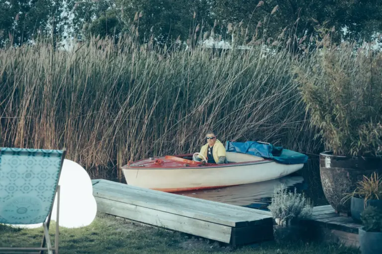 Eine Person mit Sonnenbrille sitzt in einem kleinen weißen Motorboot in der Nähe von hohem Schilf am Ufer, mit Holzplanken und Außendekoration im Vordergrund.