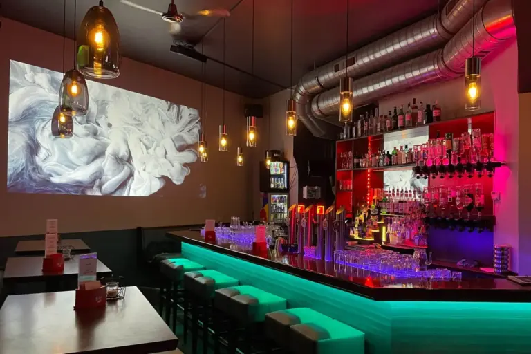 Eine moderne Bar mit leuchtenden grünen Lichtern unter dem Tresen, bunten Flaschen in Regalen, Hängelampen, Barhockern und einer Wandprojektion mit wirbelnder weißer rauchähnlicher Kunst. Die Atmosphäre ist gemütlich und einladend.