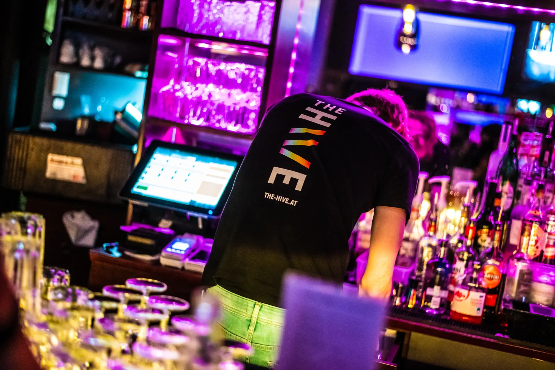 Ein Barkeeper mit einem THE THRIVE-T-Shirt arbeitet hinter einer Bar mit bunter Neonbeleuchtung, Gläsern und Flaschen, die eine lebhafte Atmosphäre im Nachtleben schaffen.