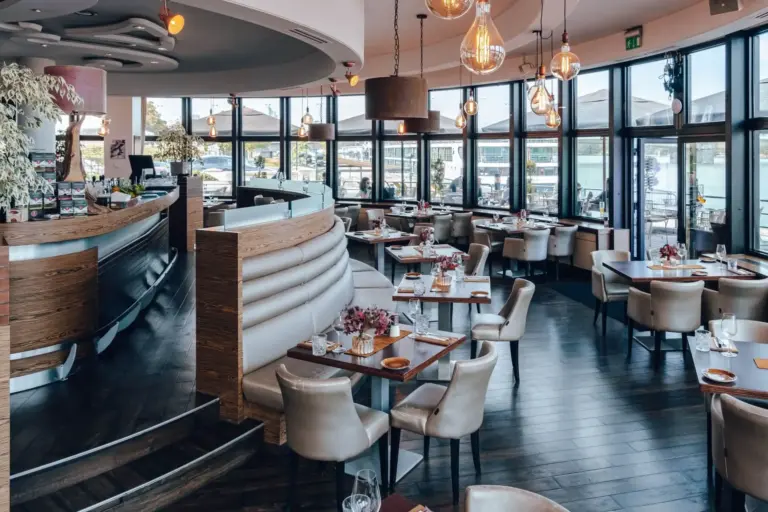 Modernes Restaurantinterieur mit geschwungenen Ständen, eleganter Hängebeleuchtung und großen Fenstern, die eine schöne Aussicht bieten. Die Tische sind ordentlich eingedeckt, und die Atmosphäre wirkt hell und einladend.