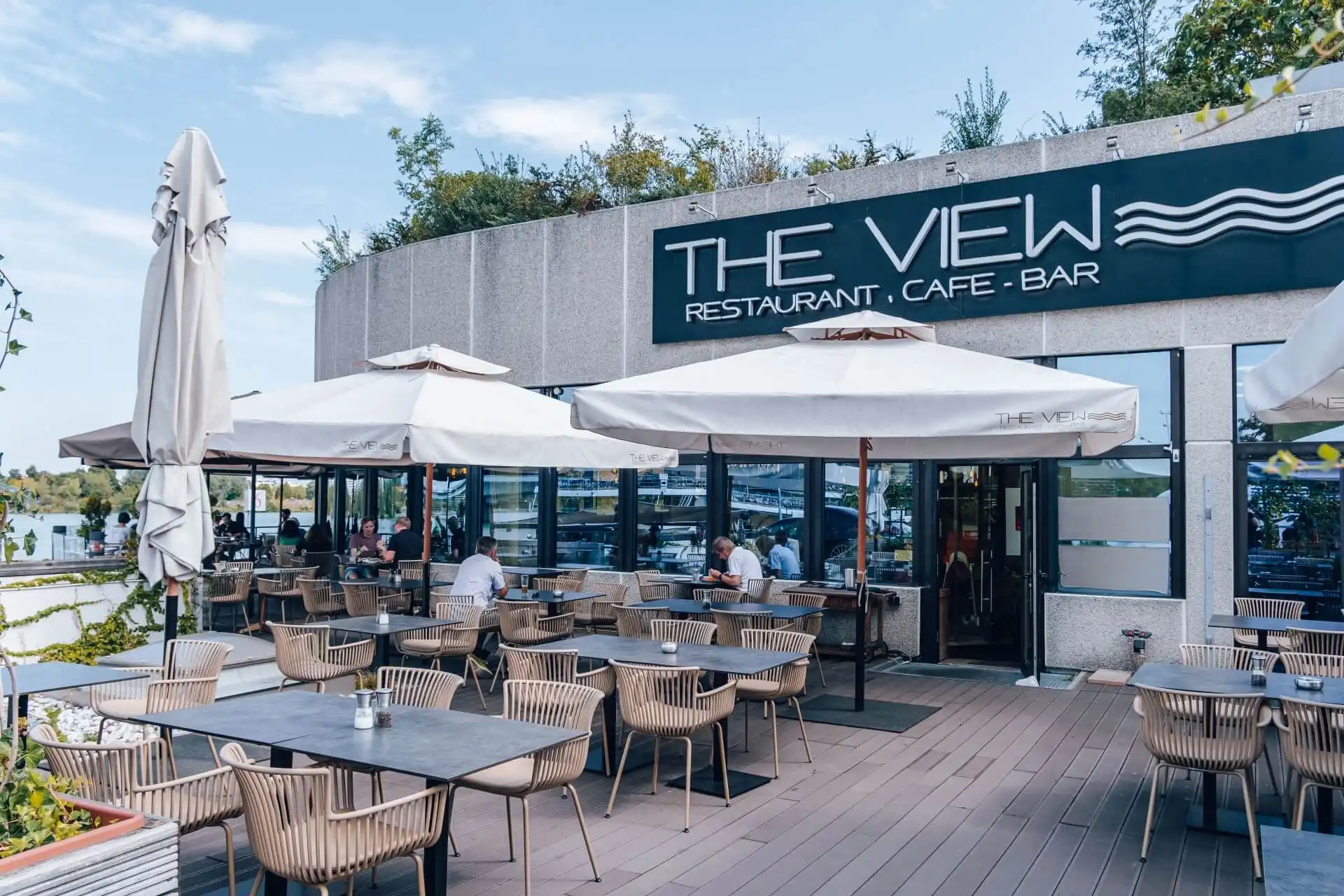 Außensitzbereich des The View Restaurant, Cafe und Bar mit Tischen, Stühlen und großen weißen Sonnenschirmen. Ein paar Leute speisen, das Gebäude hat große Fenster und ein modernes Design. Im Hintergrund sind Bäume und der Himmel zu sehen.