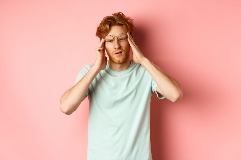 Ein Mann mit roten Haaren und Brille, der ein hellblaues T-Shirt trägt, steht vor einem rosafarbenen Hintergrund, berührt seine Schläfen und scheint Kopfschmerzen oder Kater zu haben.