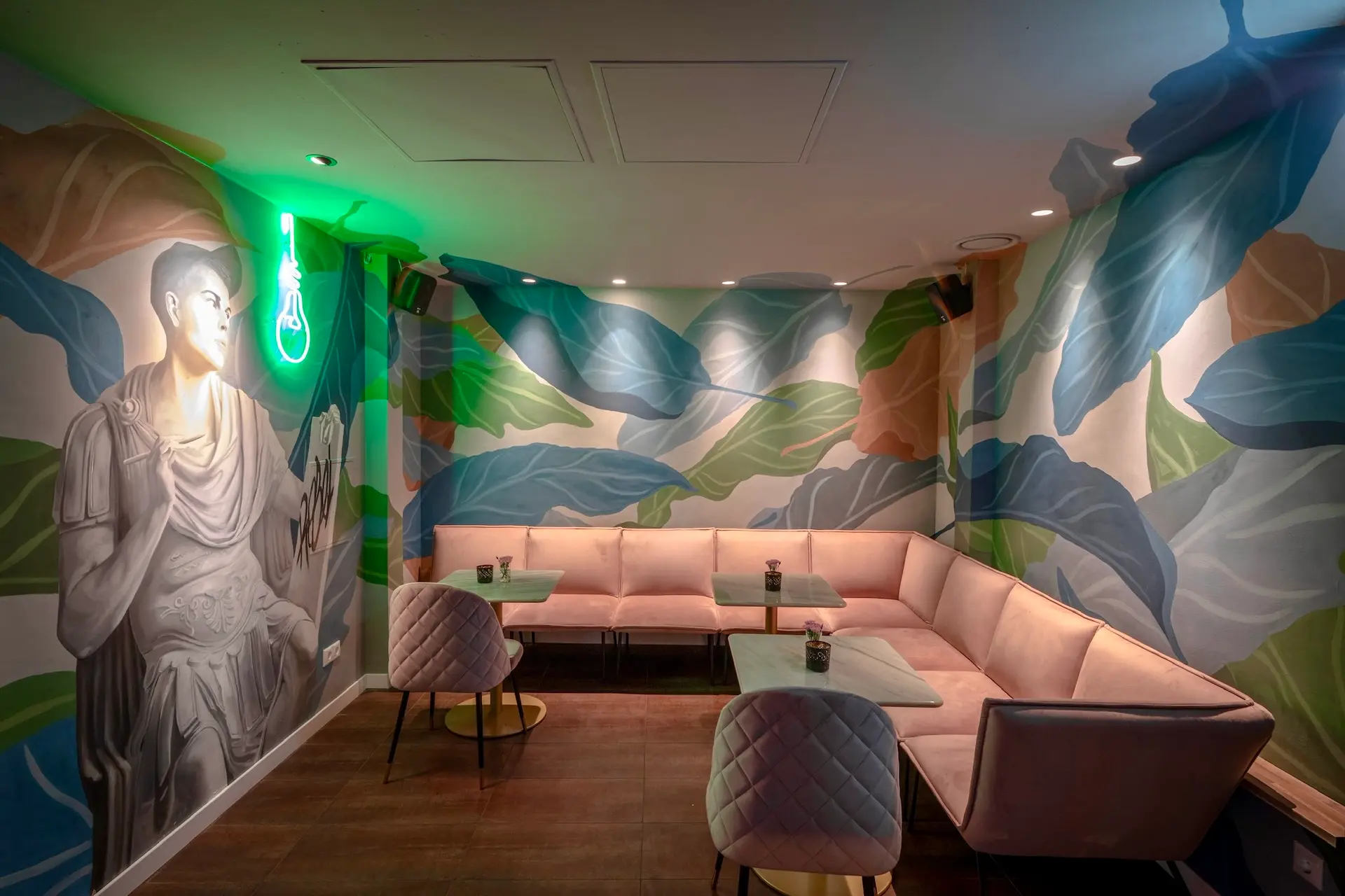 Eine stilvolle Lounge mit pastellrosa Sitzgelegenheiten, Glastischen und modernen, gesteppten Stühlen. An den Wänden befinden sich große, farbenfrohe Wandgemälde mit Blättern und eine in Graustufen gehaltene Statue mit einer neongrünen Glühbirne darüber. Die Beleuchtung ist warm und gemütlich.