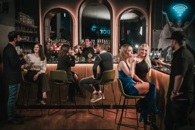 In einer stilvollen Bar mit schummriger Beleuchtung kommen die Menschen zusammen. Einige sitzen auf Barhockern, andere stehen, plaudern und trinken. An der Wand hinter dem Tresen leuchtet ein Neonschild im Toga Wien.
