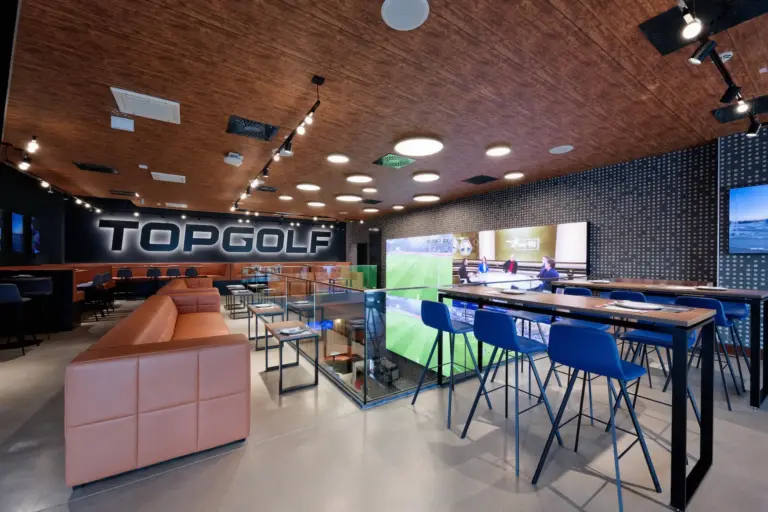 Moderne überdachte Topgolf-Lounge mit hellbraunen Sofas, blauen Hochstühlen, Tischen und Golfsimulatoren. Der Raum hat eine holzgetäfelte Decke, dunkle Akzentwände und eine große TOPGOLF-Beschilderung.