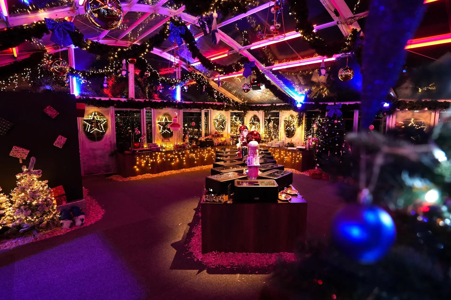 Ein festlich geschmückter Innenraum mit Weihnachtsbäumen, Girlanden, Lichterketten, Ornamenten und heller, farbenfroher Beleuchtung, die eine warme und einladende Weihnachtsatmosphäre schafft.