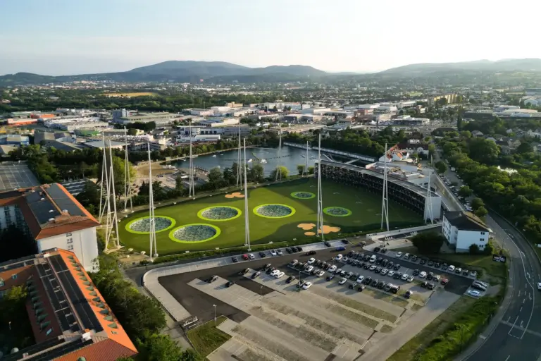 Topgolf Wien Luftaufnahme einer modernen Driving Range mit großen kreisförmigen Zielscheiben auf einer grünen Wiese, umgeben von hohen Netzen, einem Parkplatz, Gebäuden, einem Fluss und entfernten Hügeln unter einem klaren Himmel.