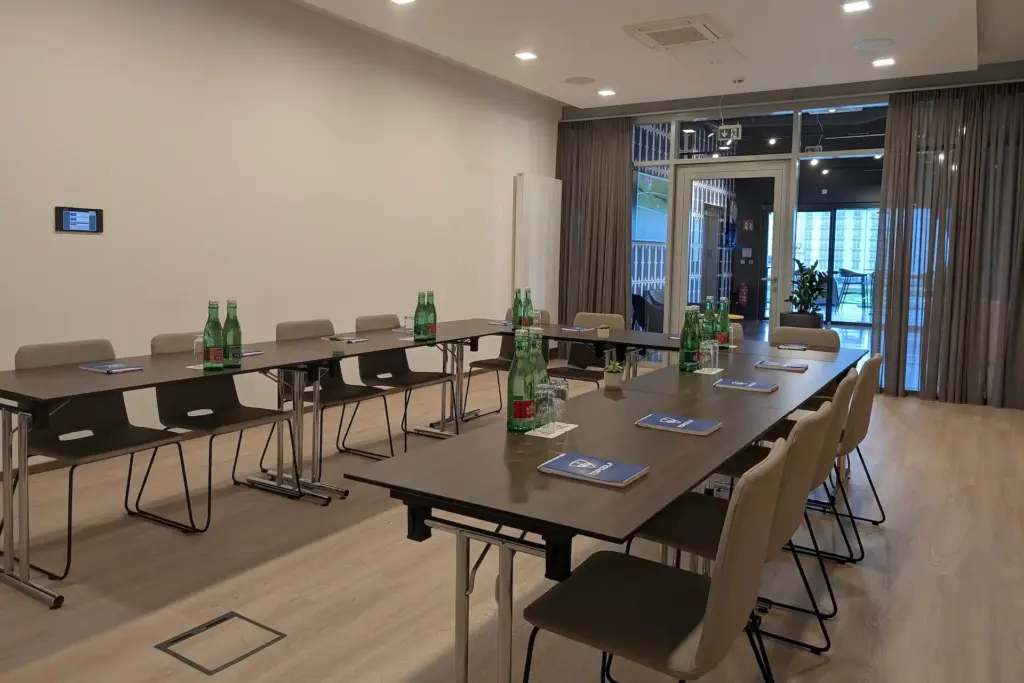 Ein moderner Konferenzraum mit zwei U-förmigen Tischen, beigen Stühlen, Wasserflaschen, Notizblöcken und Stiften, die fein säuberlich angeordnet sind. Bodentiefe Fenster mit transparenten Vorhängen lassen natürliches Licht herein.