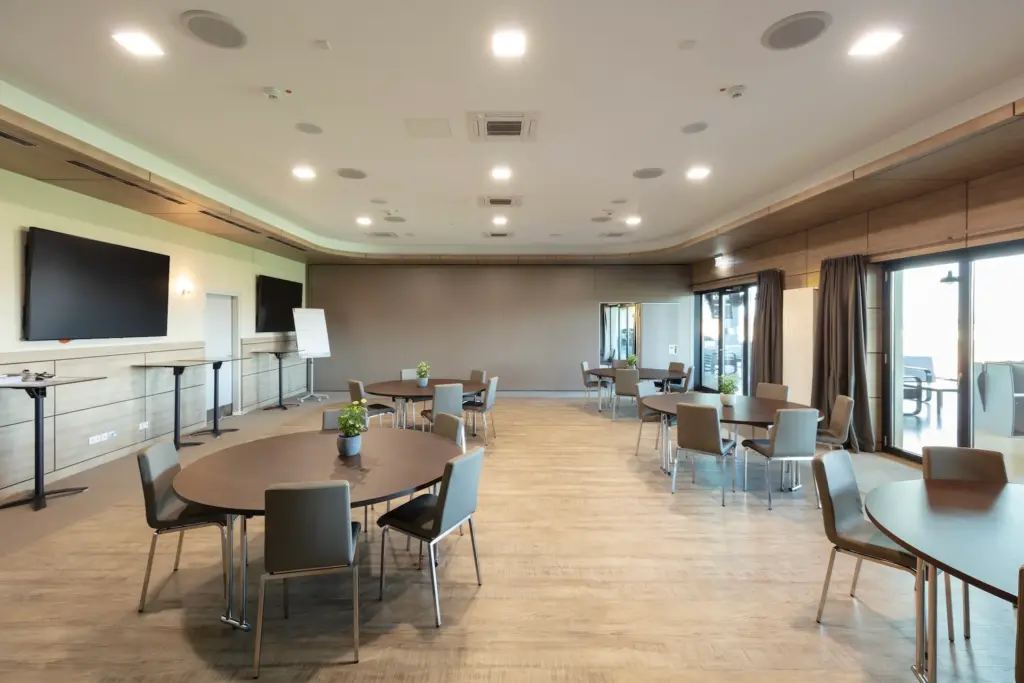 Ein geräumiger, moderner Konferenzraum mit runden Tischen und Stühlen, großen Fenstern, Flachbildfernsehern an den Wänden und hellem Holzfußboden. Auf einigen Tischen stehen kleine Pflanzen als Mittelstücke.