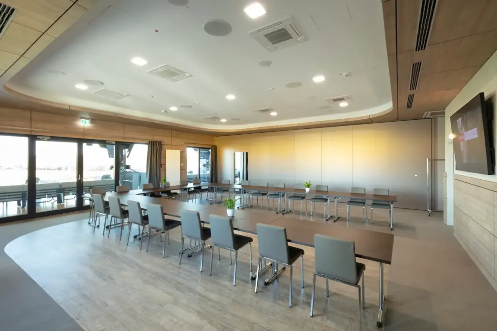 Ein moderner, geräumiger Konferenzraum mit großen Fenstern, U-förmigen Tischen, grauen Stühlen, Holzvertäfelung, Topfpflanzen auf den Tischen und einem an der Wand befestigten Bildschirm. Der Raum ist hell mit natürlichem Licht und Oberlicht.