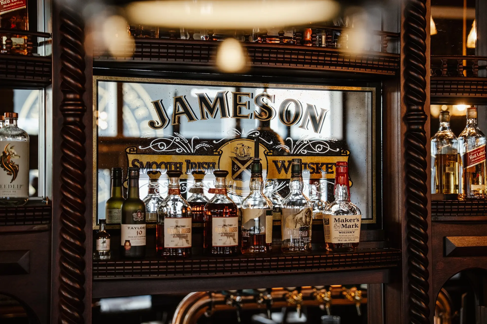 Verschiedene Spirituosenflaschen, darunter Jameson und Makers Mark, stehen in einem Barregal vor einem dekorativen Spiegel mit dem Jameson-Whiskey-Logo.