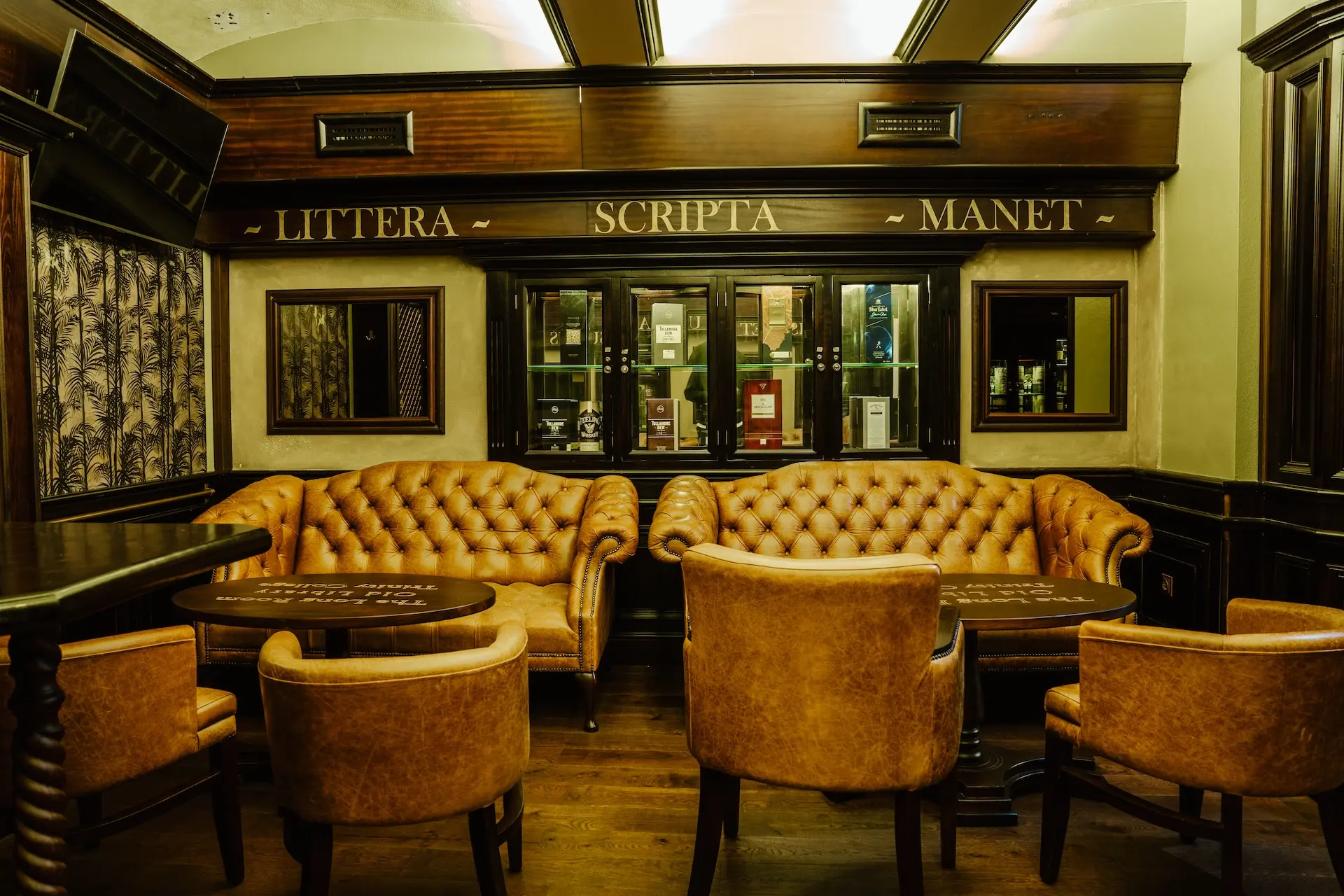 Ein gemütlicher Raum mit braunen Ledersofas und -sesseln, dunkler Holzvertäfelung und drei Spiegeln an der Wand. Über den Spiegeln stehen in goldenen Lettern die lateinischen Worte Littera Scripta Manet.