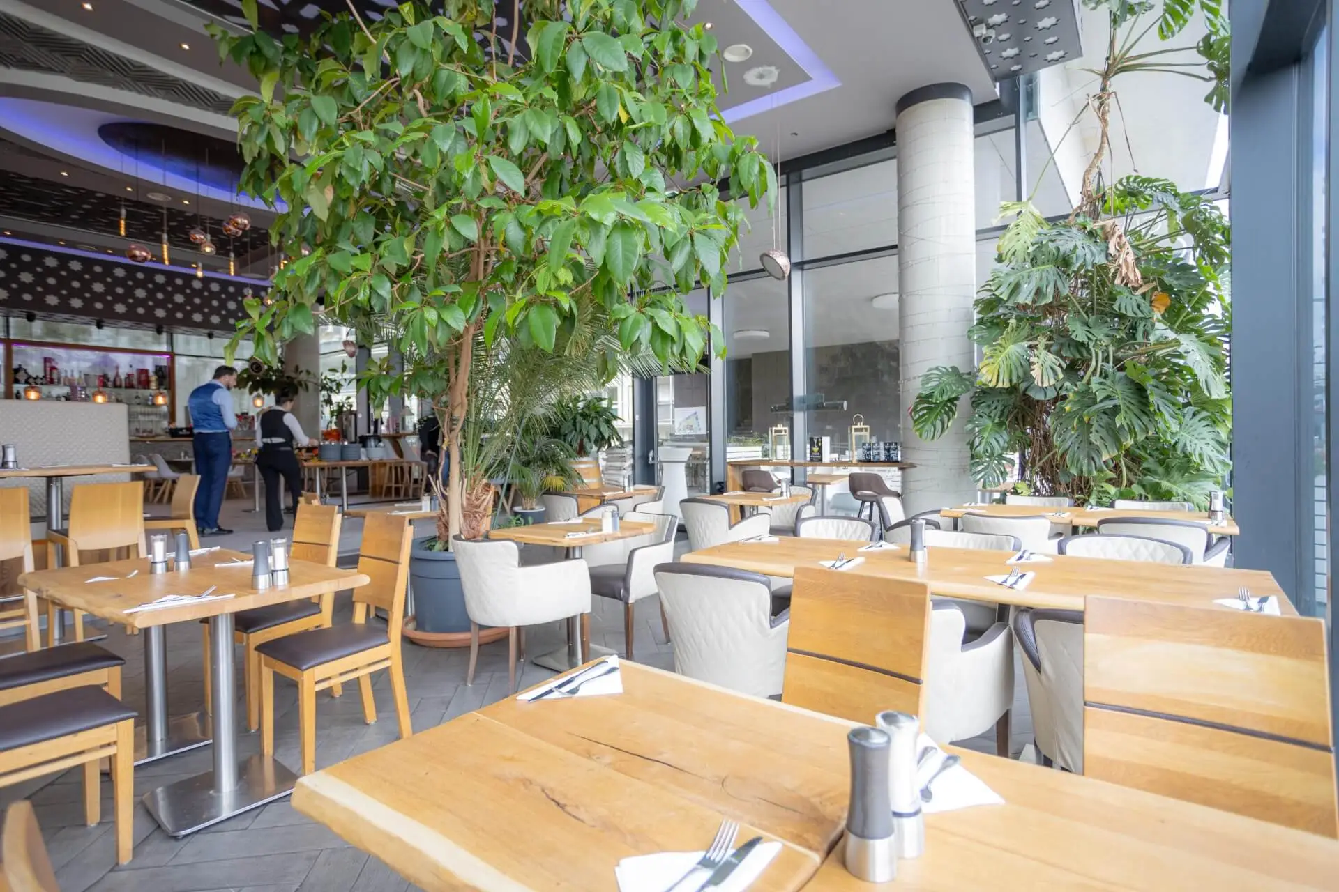 Geräumiges, modernes Restaurantinterieur mit großen Fenstern, Holztischen, gepolsterten Stühlen und üppigen Grünpflanzen. Ein paar Leute stehen im Hintergrund in der Nähe des Barbereichs, und natürliches Licht erfüllt den Raum.