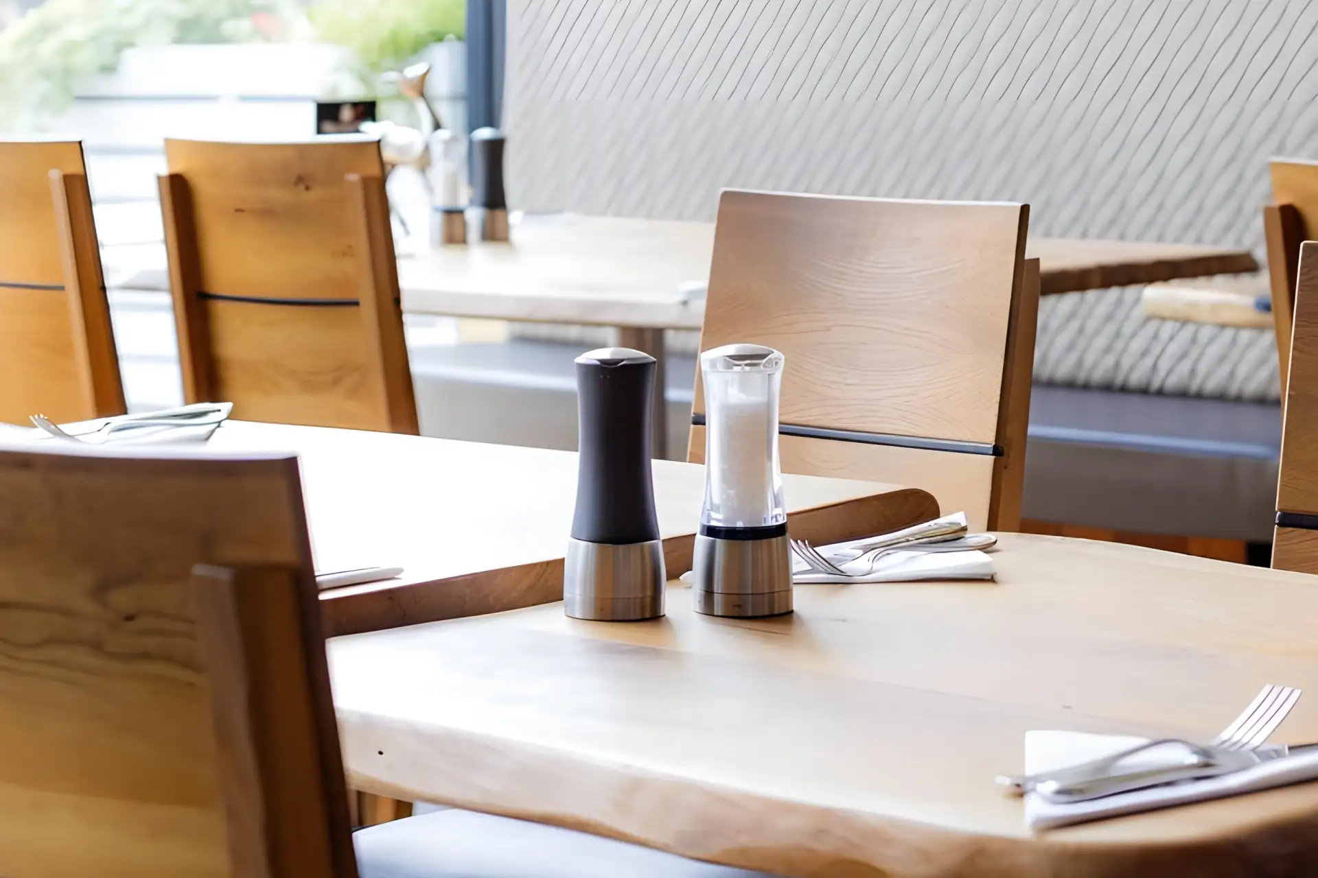 Ein modernes Restaurant mit Holztischen und -stühlen. Auf dem nächsten Tisch stehen ein schwarzes und ein durchsichtiges Salz- und Pfeffermühlenset, dazu ordentliches Besteck und Servietten.
