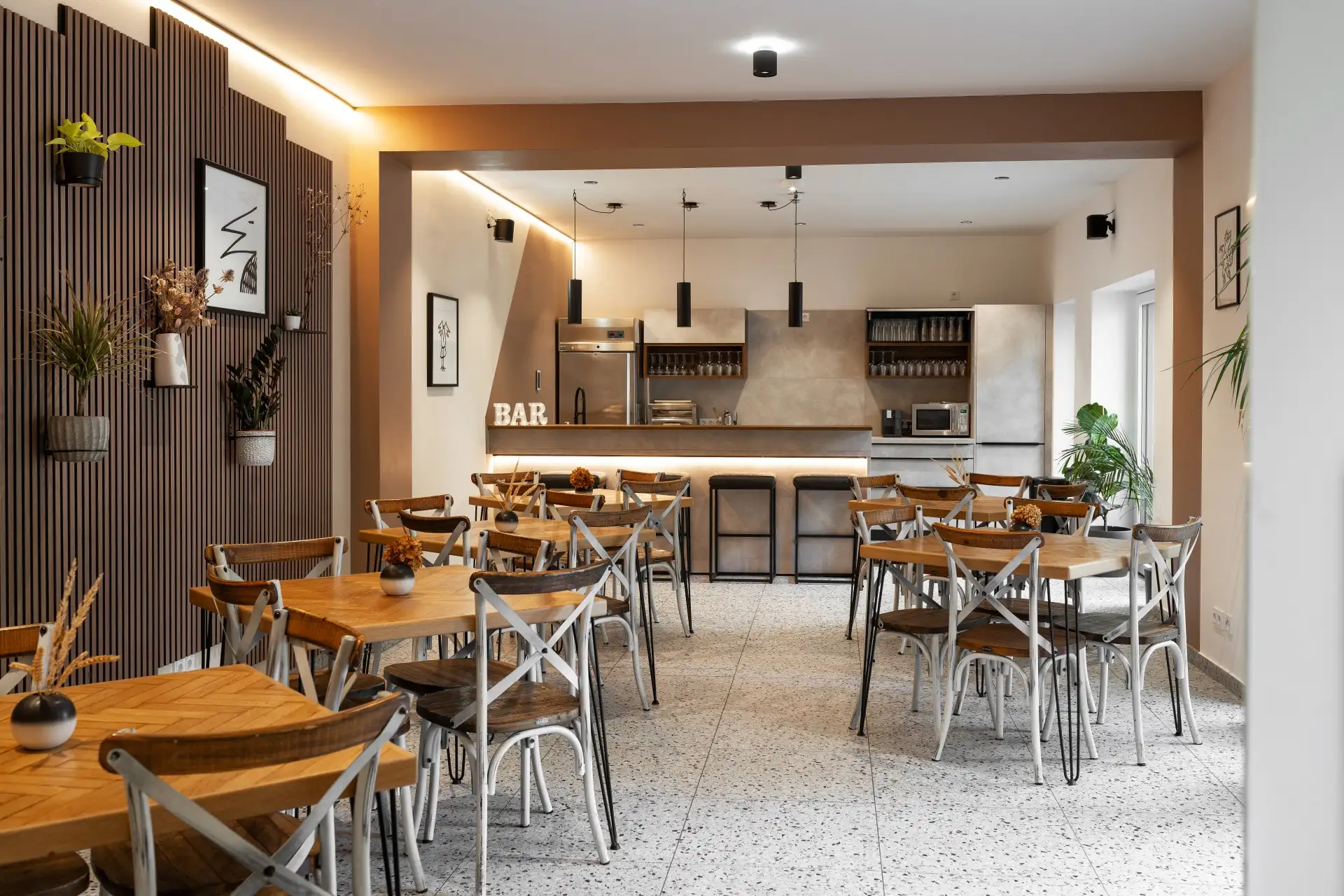 Ein modernes, gemütliches Café-Interieur mit Holztischen und -stühlen, Topfpflanzen, minimalistischer Wandkunst und einem Bartresen im Hintergrund unter Hängelampen. Der Raum ist gut beleuchtet, in neutralen Tönen gehalten und natürlich dekoriert.