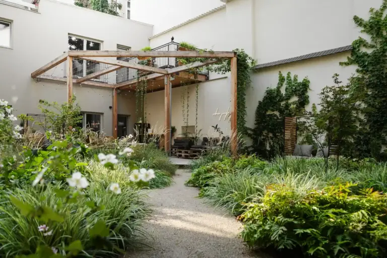 Ein üppiger Garten mit grünen Pflanzen und weißen Blumen umgibt einen Kiesweg, der zu einer überdachten Holzterrasse führt, die an ein modernes Gebäude angebaut ist, dessen weiße Wände mit Efeu bewachsen sind.