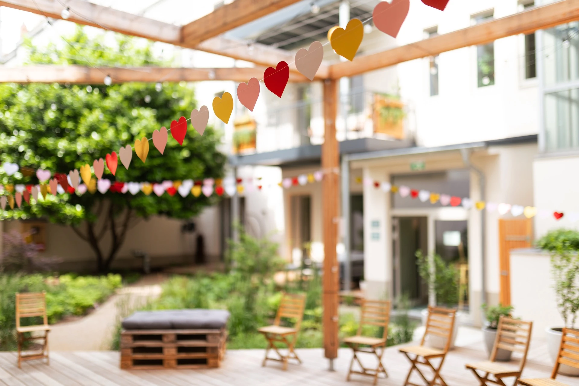 Bunte herzförmige Papiergirlanden hängen über einer Terrasse mit Holzstühlen und einer Palettenbank, umgeben von Grünzeug und einem weißen Gebäude im Hintergrund.