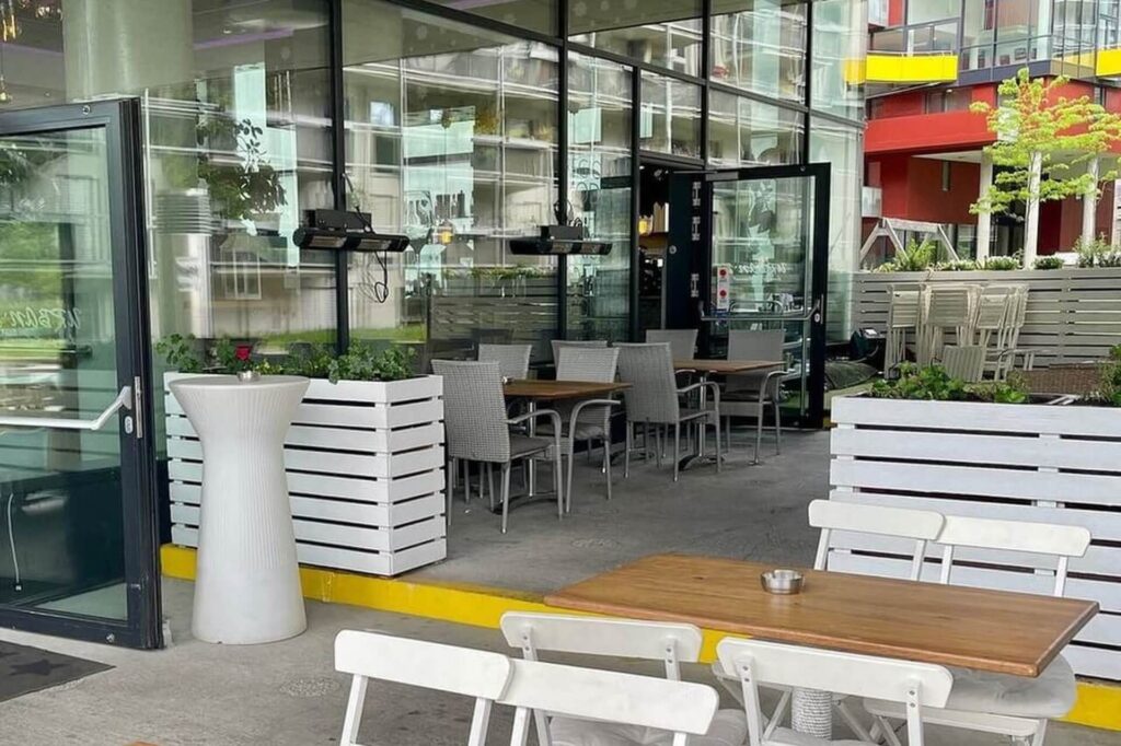 Eine moderne Schanigarten-Terrasse mit Holztischen, weißen und grauen Stühlen, weißen Pflanzkästen mit Grünzeug und Glaswänden, in denen sich die umliegenden Gebäude spiegeln. Eine offene Tür führt ins Innere des Cafés.