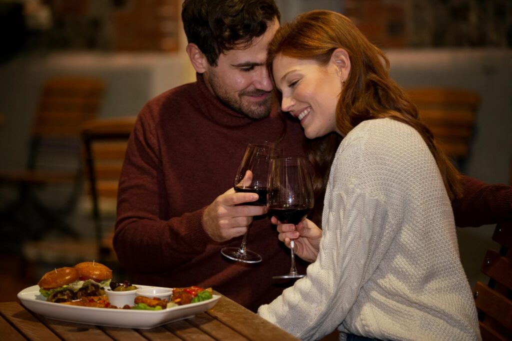 Ein lächelndes Pärchen sitzt eng beieinander an einem Tisch in Wien, hält Gläser mit Rotwein in der Hand und stößt an. Vor ihnen steht ein Teller mit Burgern und Beilagen, der eine gemütliche und intime Valentinstagsatmosphäre schafft.