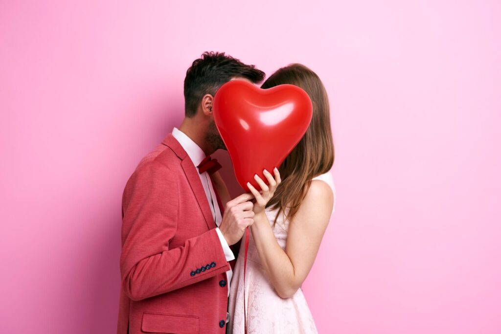 In dieser charmanten Szene zum Valentinstag in Wien steht ein Paar vor einem rosafarbenen Hintergrund dicht beieinander und hält einen roten herzförmigen Ballon, der ihre Gesichter bedeckt, während sie sich zu küssen oder zu umarmen scheinen.