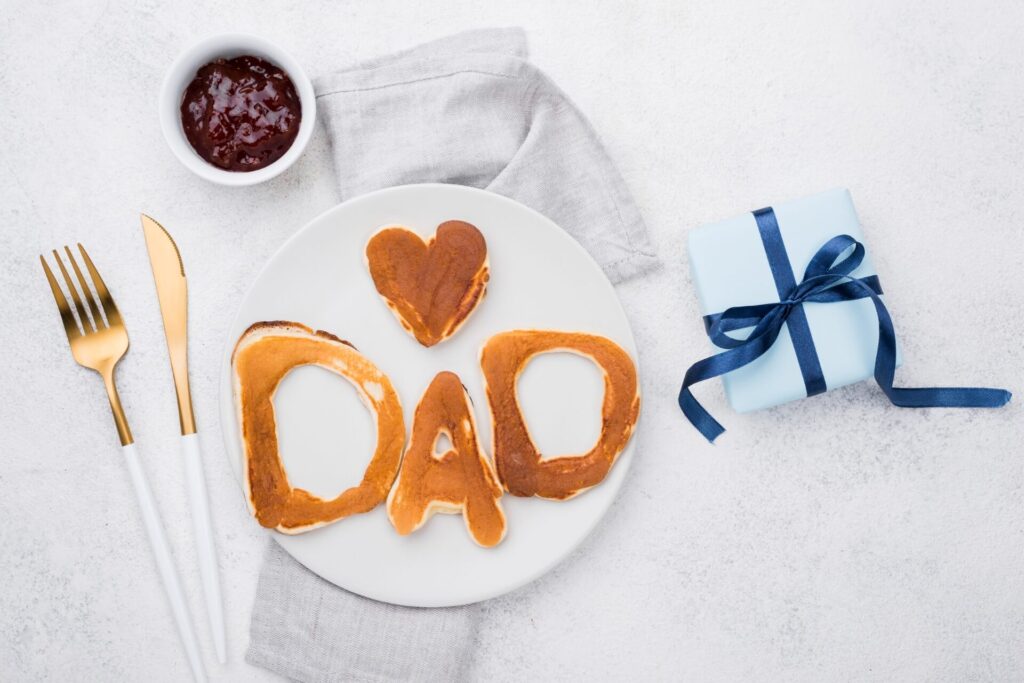 Ein Teller mit Pfannkuchen in Form von DAD und einem Herz, daneben eine goldene Gabel und ein goldenes Messer, eine kleine Schale mit Marmelade, eine graue Serviette und eine blaue Geschenkbox mit einem marineblauen Band - perfekt für den Vatertag in Wien.