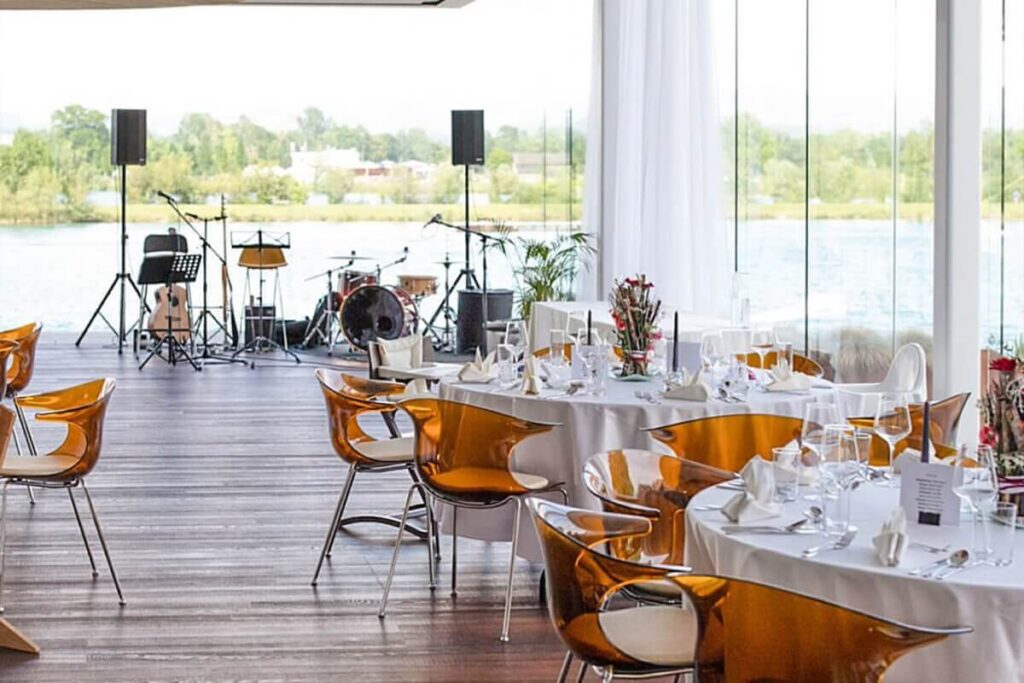Ein moderner Veranstaltungsraum mit runden Tischen zum Essen, bernsteinfarbenen Stühlen und großen Glasfenstern mit Blick aufs Wasser. Im Hintergrund ist eine Bühne mit Musikinstrumenten und Lautsprechern aufgebaut.