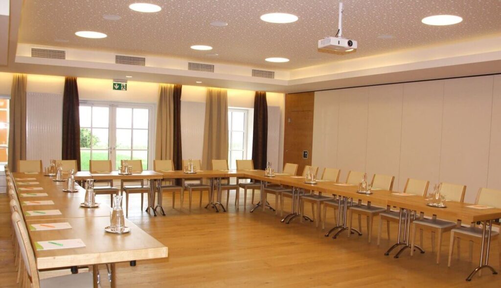 Ein moderner Veranstaltungsraum mit U-förmigen Holztischen, beigen Stühlen, Wasserkaraffen, Notizblöcken und Gläsern, bereit für eine Besprechung. Große Fenster füllen den Raum mit natürlichem Licht, während an der Decke ein Projektor angebracht ist.