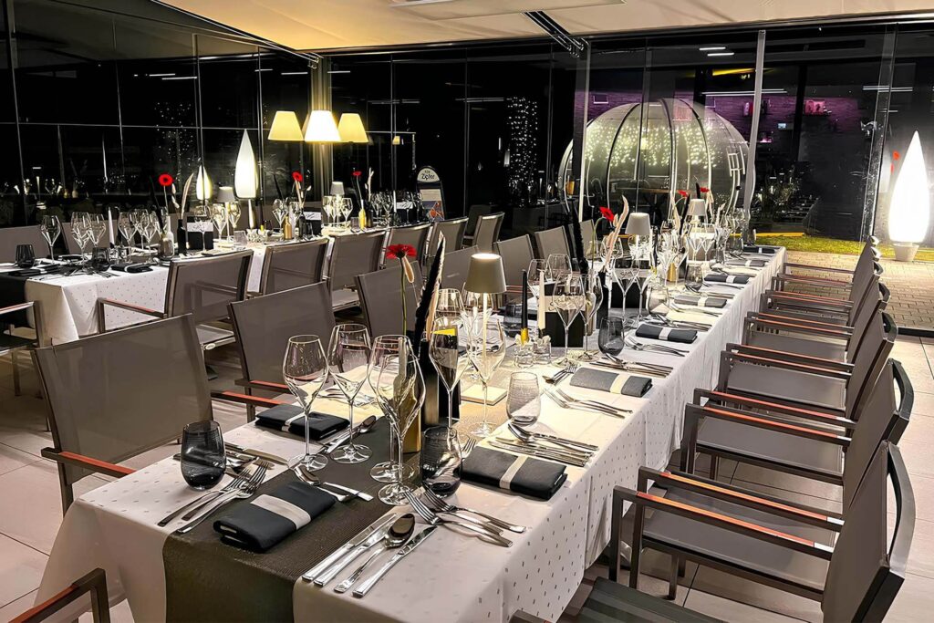 Für ein formelles Abendessen im Veranstaltungsraum sind elegante Esstische mit weißen Tischtüchern, gefalteten Servietten, Weingläsern und Besteck gedeckt. Rote Blumen schmücken jedes Gedeck. Große Fenster geben den Blick frei auf einen beleuchteten Außenbereich mit Kuppelstrukturen.
