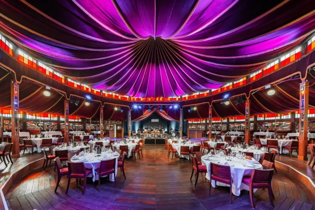 Eleganter Speisesaal mit runden Tischen und roten Stühlen auf dunklem Holzboden, unter einer großen violetten Zeltdecke mit warmer Beleuchtung und bunten Glasmalereien, mit Blick auf eine Bühne - ein außergewöhnlicher Veranstaltungssaal in Wien.