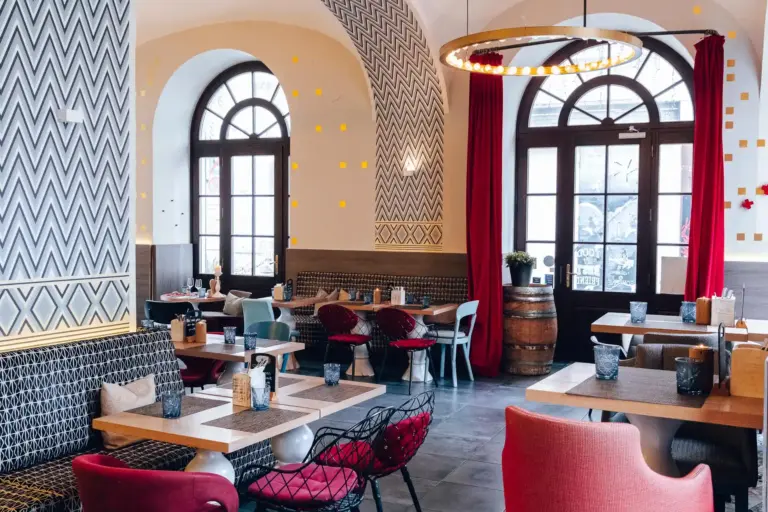 Ein modernes, stilvolles Restaurant mit gemusterten Wänden, gewölbten Fenstern, roten Vorhängen, Holztischen und einer Mischung aus bunten Stühlen. Die Tische sind mit Gläsern und Servietten gedeckt, und der Raum ist von natürlichem Licht durchflutet.