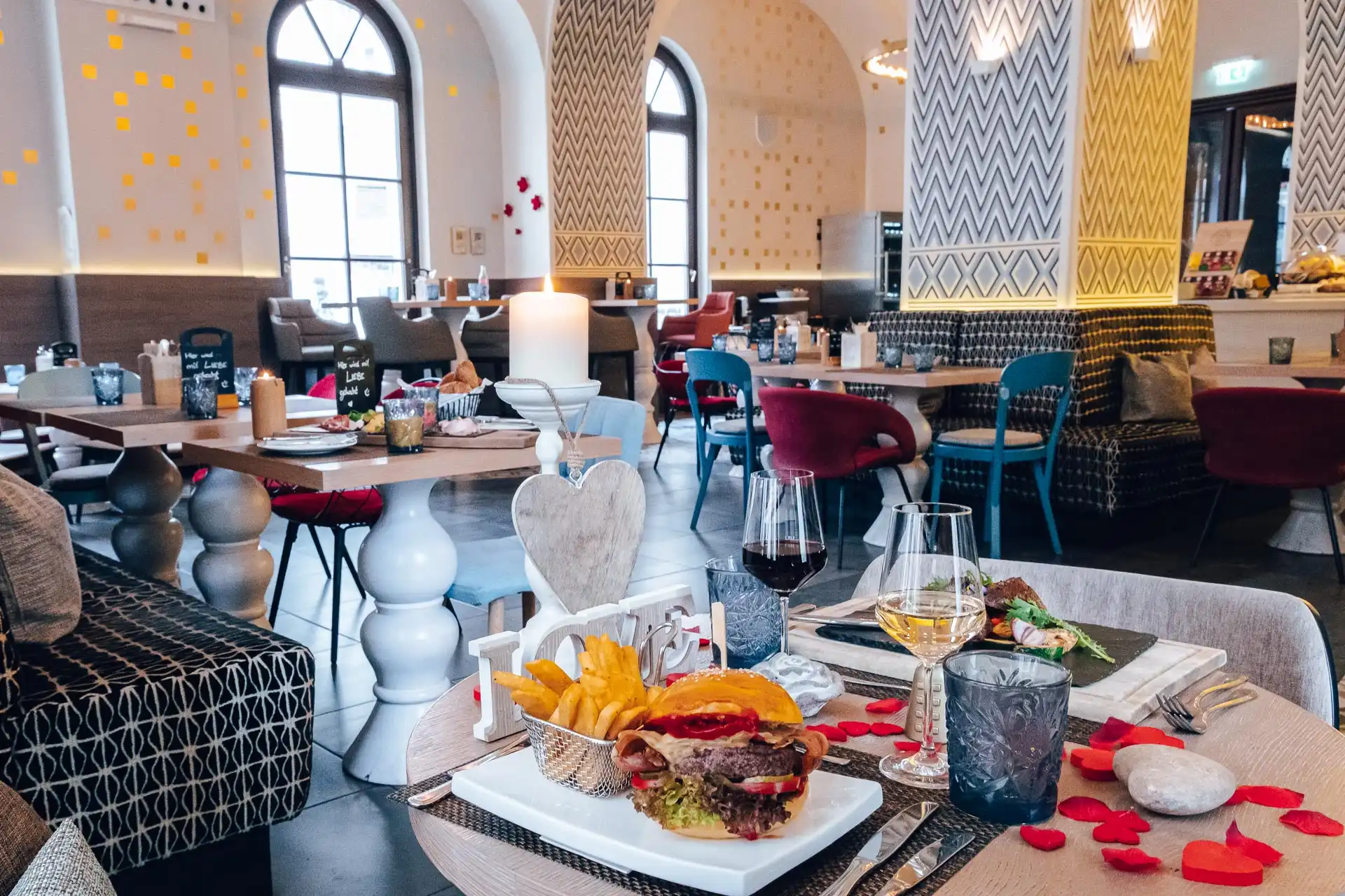 Ein stilvolles Restaurantinterieur mit elegantem Dekor, gedeckten Tischen und natürlichem Licht aus hohen Fenstern. Im Vordergrund ist ein Tisch mit Burgern, Pommes frites, Getränken und verstreuten Rosenblättern gedeckt, was eine romantische Atmosphäre schafft.