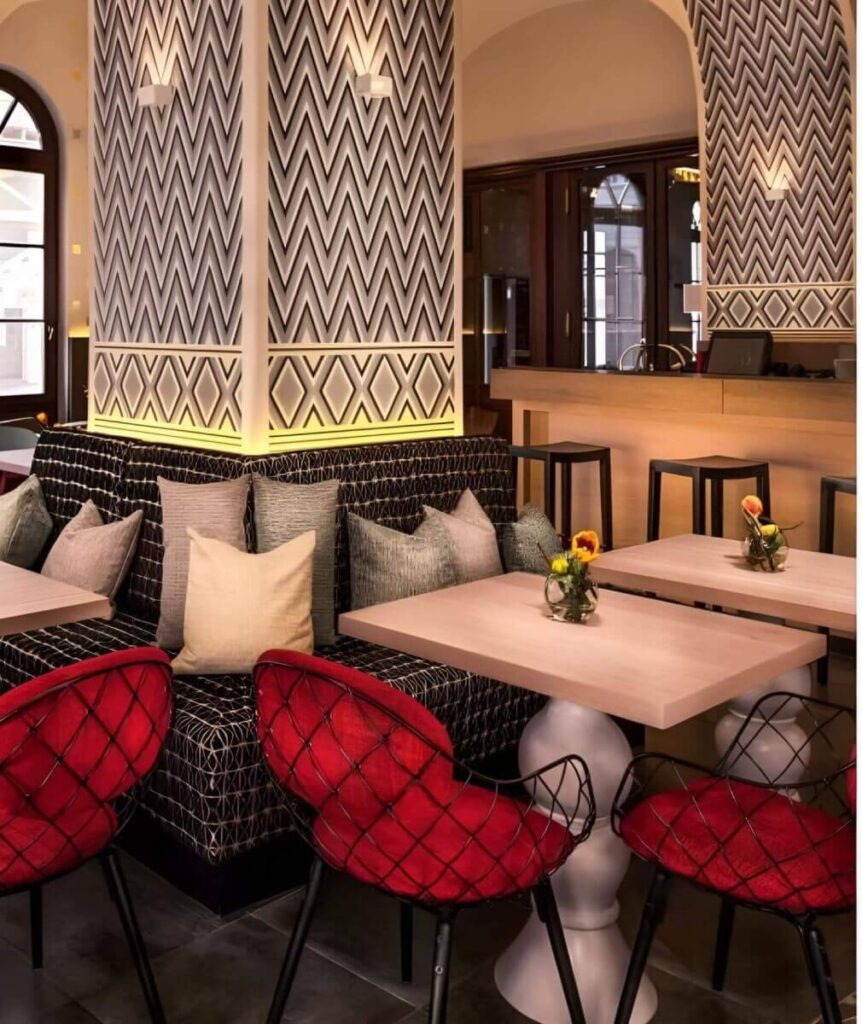 Modernes Café-Interieur mit geometrisch gemusterten Wänden, gepolsterten Sitzecken und roten Drahtstühlen. Helle Holztische mit kleinen Vasen mit gelben Blumen schaffen einen gemütlichen, stilvollen Raum - perfekt, wenn Sie den Partyraum mieten Wien für Ihre nächste Zusammenkunft nutzen möchten.