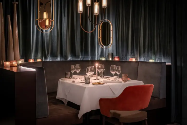 Ein schummriges, elegantes Restaurant mit weißem Tischtuch, gedeckt für vier Personen mit Weingläsern und Kerzen. Samtvorhänge, moderne Pendelleuchten und Spiegel schaffen eine gemütliche, gehobene Atmosphäre.