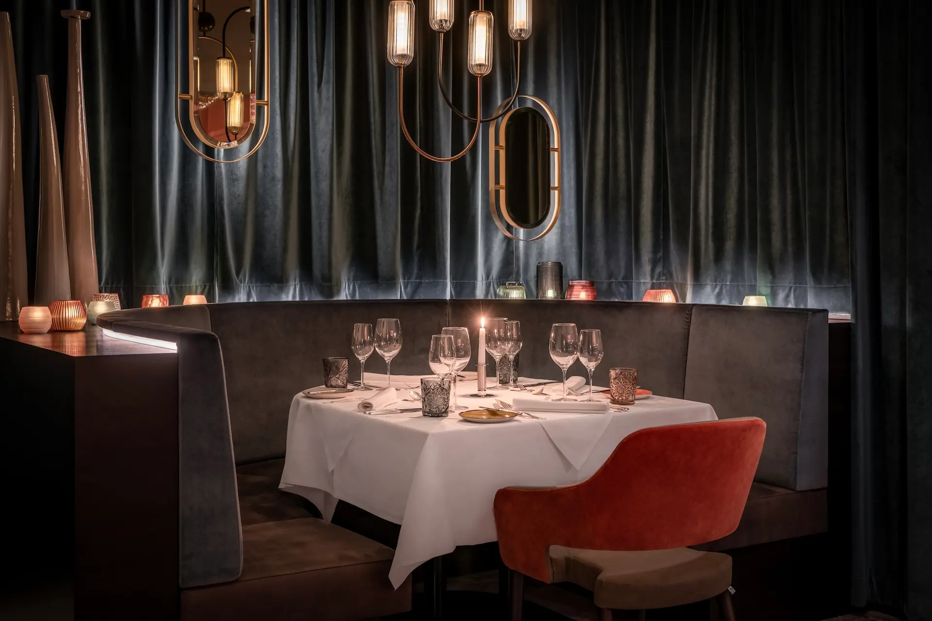 Ein schummriges, elegantes Restaurant mit weißem Tischtuch, gedeckt für vier Personen mit Weingläsern und Kerzen. Samtvorhänge, moderne Pendelleuchten und Spiegel schaffen eine gemütliche, gehobene Atmosphäre.
