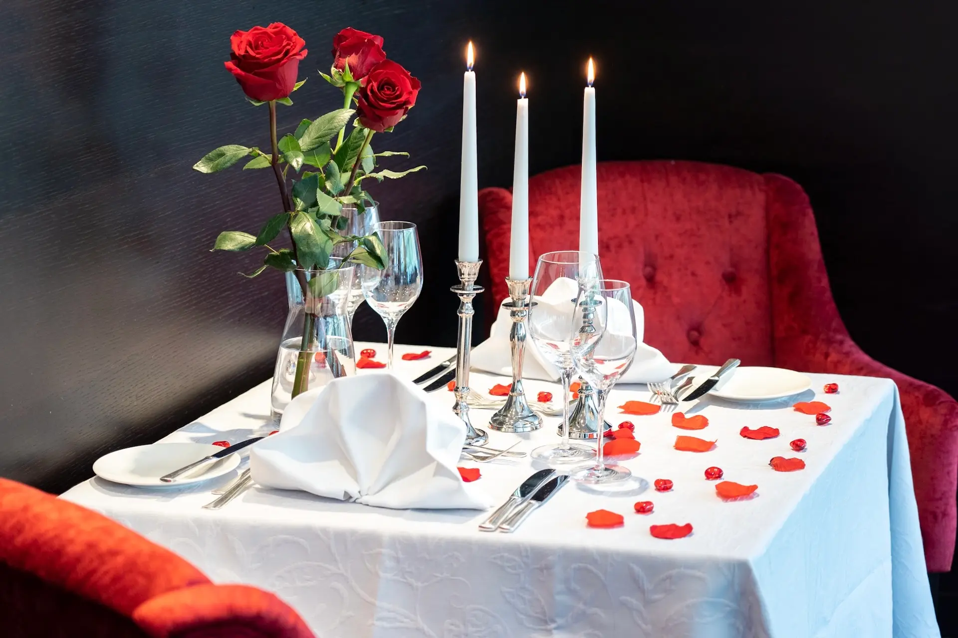 Ein romantisch gedeckter Tisch mit zwei roten Stühlen, einer weißen Tischdecke, drei brennenden Kerzen, roten Rosenblättern und einer Vase mit roten Rosen, gedeckt für zwei Personen. Weingläser und Silberbesteck sind ordentlich angeordnet.