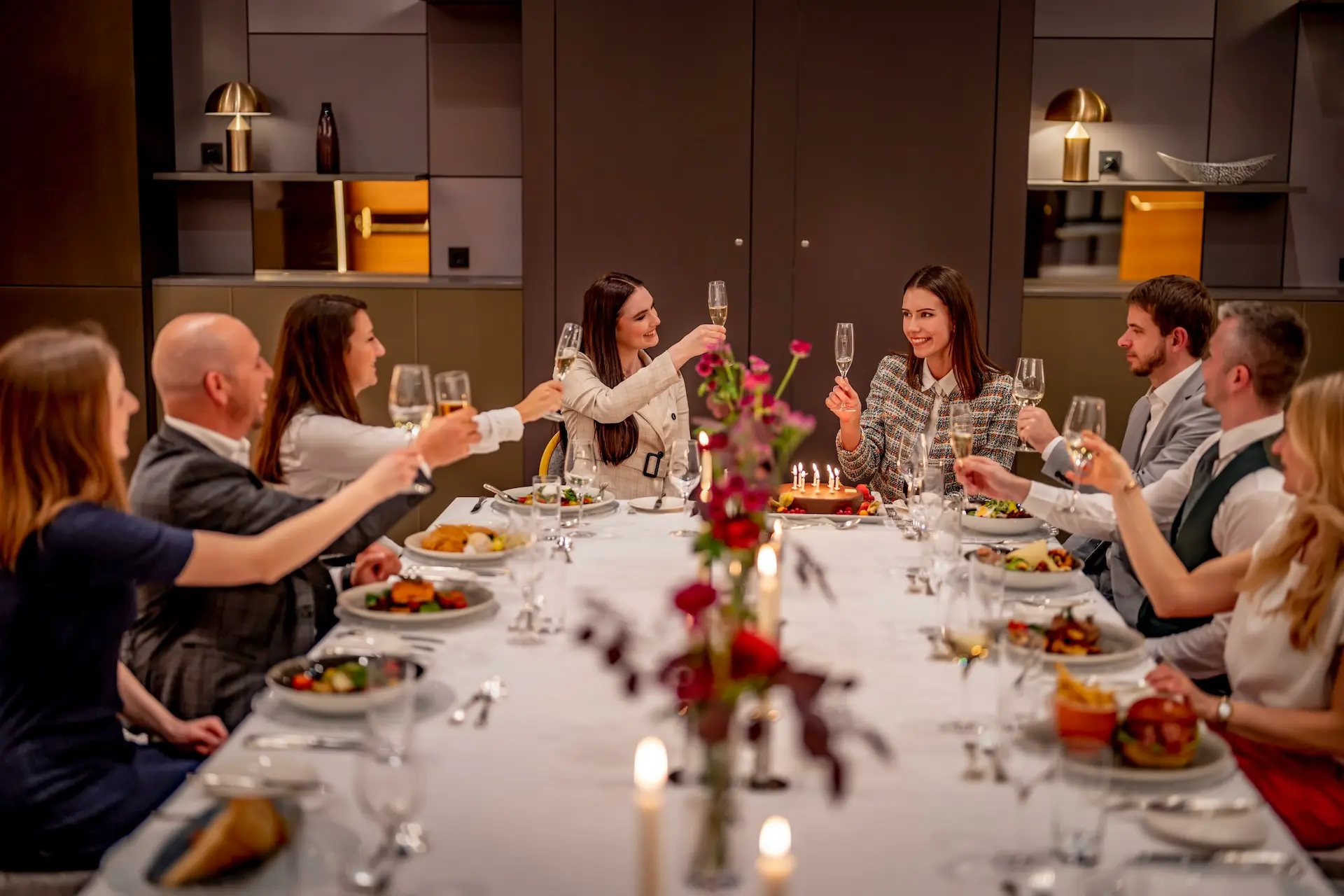 Eine Gruppe von sieben Personen sitzt um einen Tisch herum, stößt mit Gläsern an und feiert mit Essen und einem Kuchen mit Kerzen in der Mitte des Tisches. Die Atmosphäre ist festlich und warm.