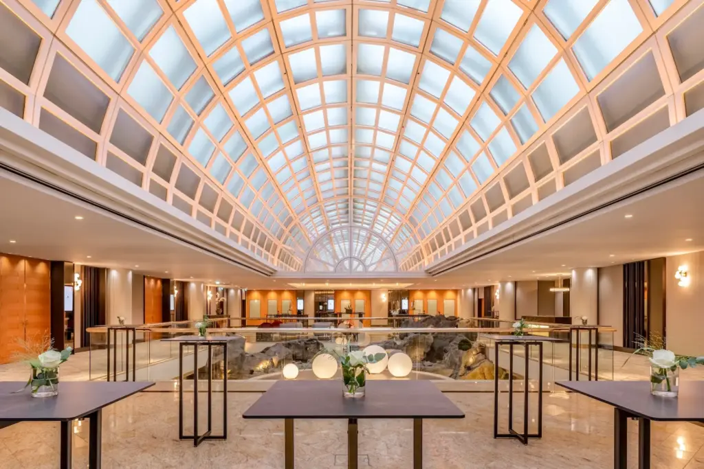 Ein geräumiger, moderner Saal mit einer hohen, gewölbten Glasdecke, Marmorböden und hohen, mit kleinen Blumenarrangements geschmückten Tischen. Kreisförmige Lichter sind um ein felsiges Mittelstück in der Mitte angeordnet.