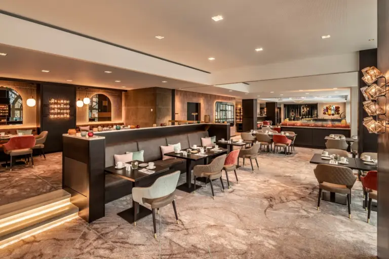 Modernes Restaurantinterieur mit gepolsterten Stühlen, gedeckten Tischen, gedämpfter Beleuchtung und zeitgenössischem Dekor. Erdtöne und stilvolle Akzente schaffen eine warme, einladende Atmosphäre mit Sitzgelegenheiten an Tischen und Stühlen.
