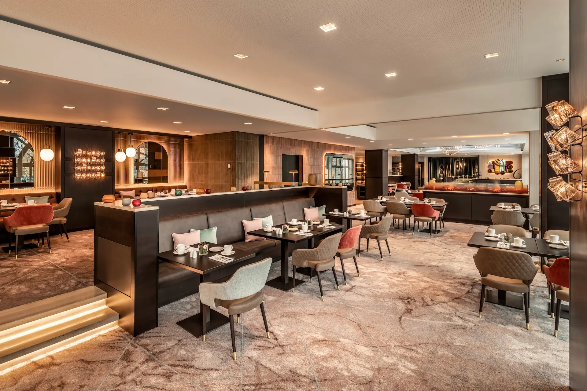 Modernes Restaurantinterieur mit gepolsterten Stühlen, gedeckten Tischen, gedämpfter Beleuchtung und zeitgenössischem Dekor. Erdtöne und stilvolle Akzente schaffen eine warme, einladende Atmosphäre mit Sitzgelegenheiten an Tischen und Stühlen.