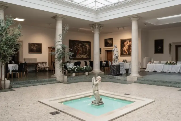Eine Marmorstatue steht in der Mitte eines kleinen Zimmerbrunnens, umgeben von hohen Säulen, klassischen Skulpturen, Esstischen und Gemälden in einer eleganten, gut beleuchteten Kunstgalerie oder einem Bankettsaal.