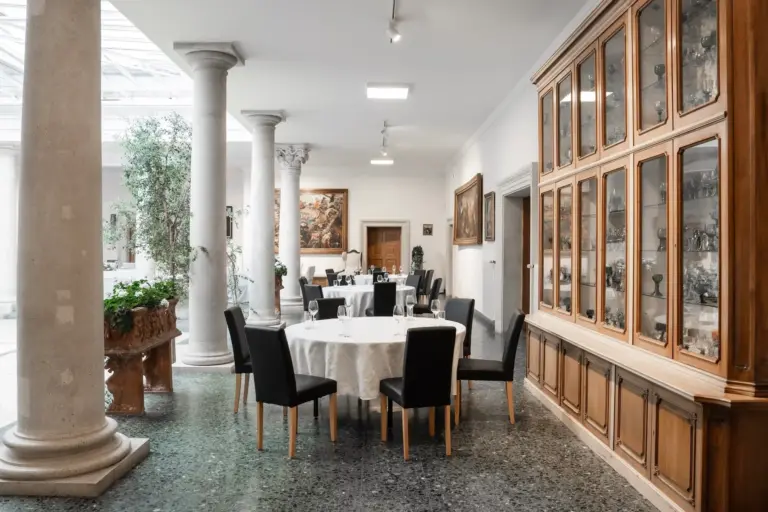 Elegantes Esszimmer mit runden Tischen, die für eine Mahlzeit gedeckt sind, umgeben von schwarzen Stühlen. An den Wänden stehen Glasvitrinen, die mit Gegenständen gefüllt sind, und große Säulen und Pflanzen fügen klassische Dekorationselemente hinzu. An den weißen Wänden hängen Gemälde.