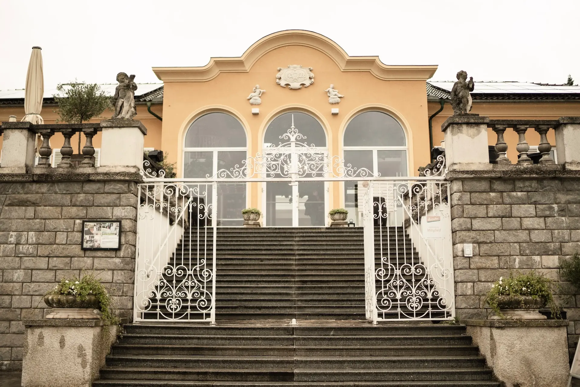 Eine große Steintreppe mit verziertem weißem Geländer führt zu einem pfirsichfarbenen Gebäude mit großen Rundbogenfenstern. Auf beiden Seiten der Treppe stehen Steinstatuen, und Topfpflanzen schmücken den Eingang.
