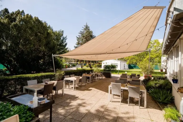 Volee Terrasse im Freien mit Tischen und Stühlen unter einem großen, beigen, dreieckigen Sonnenschirm, umgeben von Grünflächen und Bäumen. Der Bereich ist sonnig, und über den Köpfen ist der blaue Himmel zu sehen.