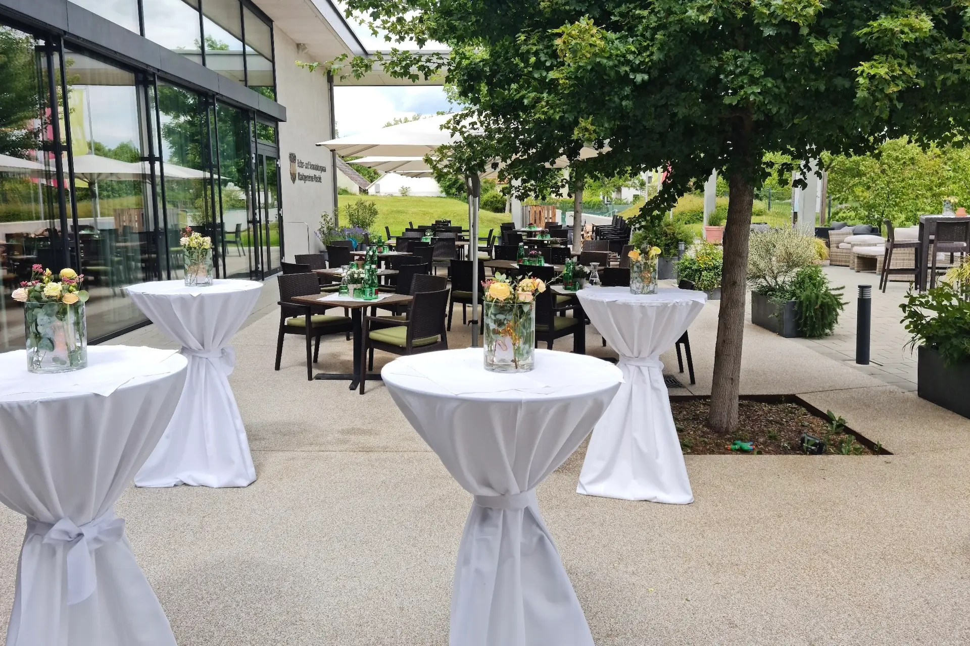Außenterrasse mit mehreren mit weißen Tüchern bedeckten Cocktailtischen, auf denen jeweils eine Glasvase mit Blumen steht, umgeben von Esstischen und Stühlen, beschattet von Bäumen und großen Sonnenschirmen in der Nähe eines modernen Gebäudes.