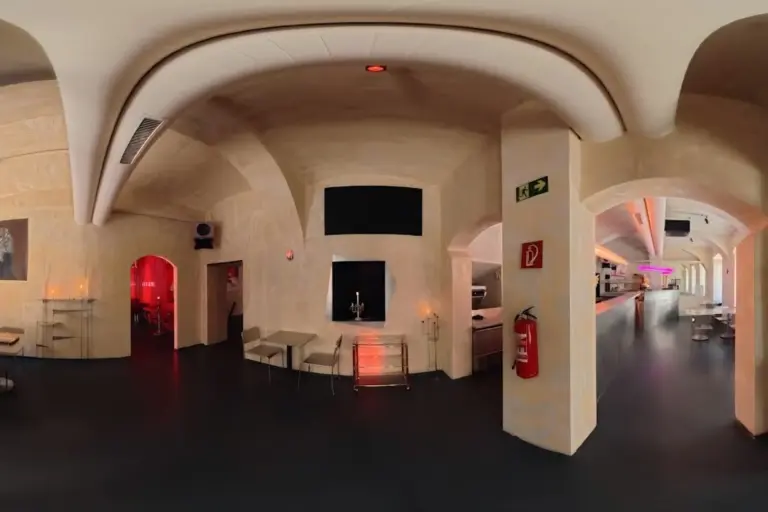 Eine moderne, minimalistische Bar mit gewölbten Decken, cremefarbenen Wänden, dezenter orange- und rosafarbener Beleuchtung, einem langen Tresen und verstreuten Tischen und Stühlen an den Wänden.