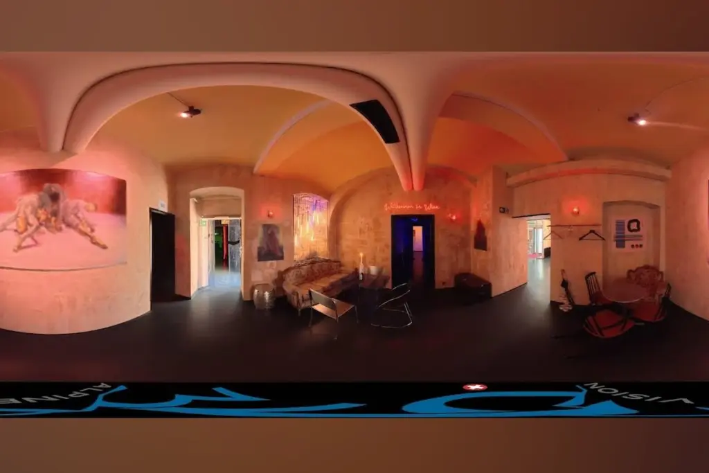 Eine 360-Grad-Ansicht eines Kunstateliers mit gewölbten Decken, warmer Beleuchtung, Gemälden an den Wänden, einem Sofa, Stühlen, einer Garderobe und einer Tür, die in einen anderen Raum führt. Der Raum hat eine gemütliche, kreative Atmosphäre.