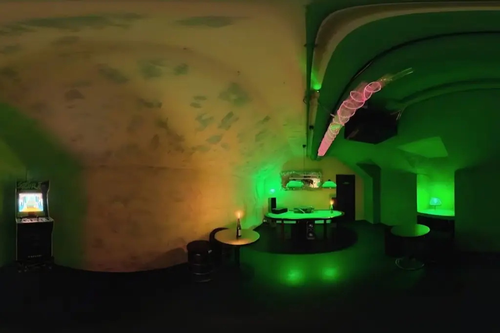 Ein schwach beleuchteter Raum mit grüner Beleuchtung, einer gewölbten Decke, runden Tischen, einem Schreibtisch mit Geräten, einem Spielautomaten und von der Decke hängenden dekorativen Lampen.