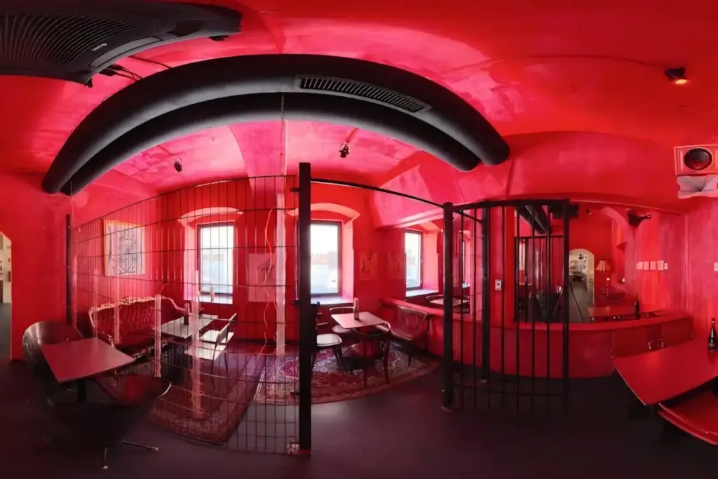 Weltraum Bar Ein Panoramablick auf einen leuchtend roten Raum mit vergitterten Toren, modernen Möbeln und drei Fenstern. Der Raum verfügt über gebogene schwarze Deckenrohre, Tische, Stühle, einen Teppich und eine eklektische Einrichtung, die ihm ein industrielles, eklektisches Flair verleiht.