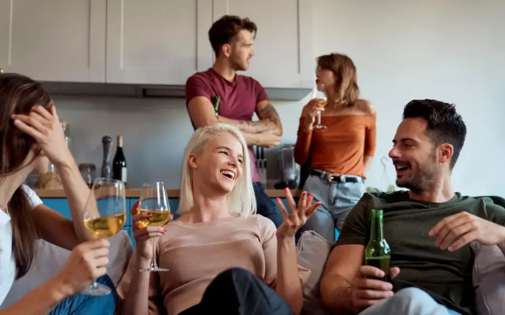 Fünf junge Erwachsene sitzen in einer modernen Küche und einem Wohnbereich, lachen und unterhalten sich mit Getränken in der Hand, während sie gemeinsam Wahrheit oder Pflicht Fragen genießen. Zwei stehen im Hintergrund, während drei sich auf einer Couch im Vordergrund entspannen.