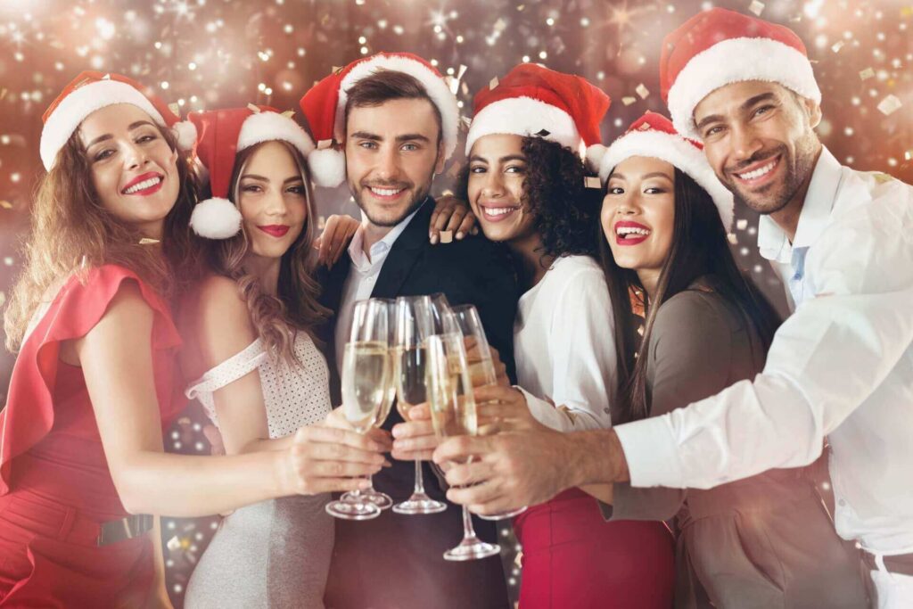 Sechs Personen mit Weihnachtsmannmützen und festlichen Kostümen lächeln und stoßen mit Sektgläsern auf einer Weihnachtsfeier in Wien an, während im Hintergrund funkelnde Lichter funkeln.